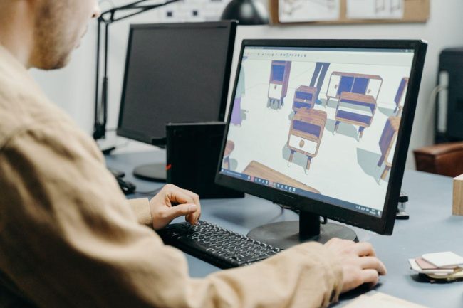Designer produit travaillant sur un logiciel de modélisation 3D affichant des fauteuils à l'écran, dans un bureau équipé de plusieurs moniteurs